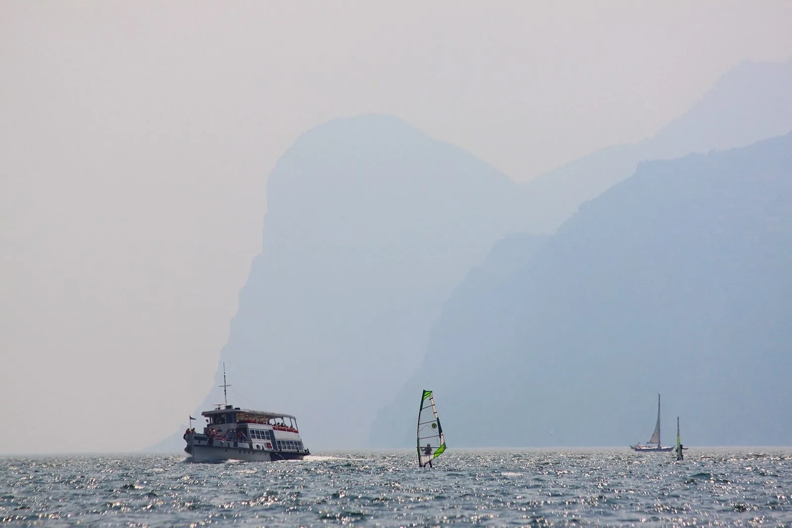 Un battello di linea naviga sul Lago di Garda tra windsurf e barche a vela, con le montagne dell'Alto Garda nella foschia